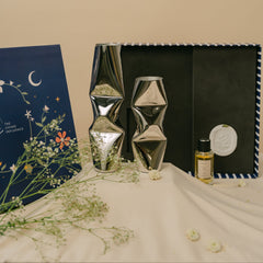 Two metallic silver geometric Chrome vases alongside a small bottle of room perfume with Diffusion Stone displayed next to a navy blue gift box.