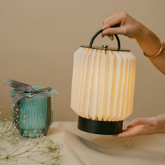 A hand-held portable pleated lantern lamp with a warm glowing cream shade and black base/handle along with a small turquoise votive candle with a gray ribbon.