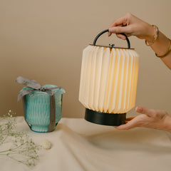 A hand-held pleated lantern-style portable lamp with a warm glowing white shade, along with Flower Cut Votive Candle wrapped like a gift