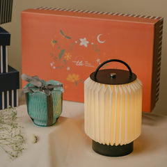 A ribbed white pleated portable table lamp with a black handle and base, shown next to a small teal ribbed Flower Cut Votive Candle and a large orange decorative gift box on a tabletop.