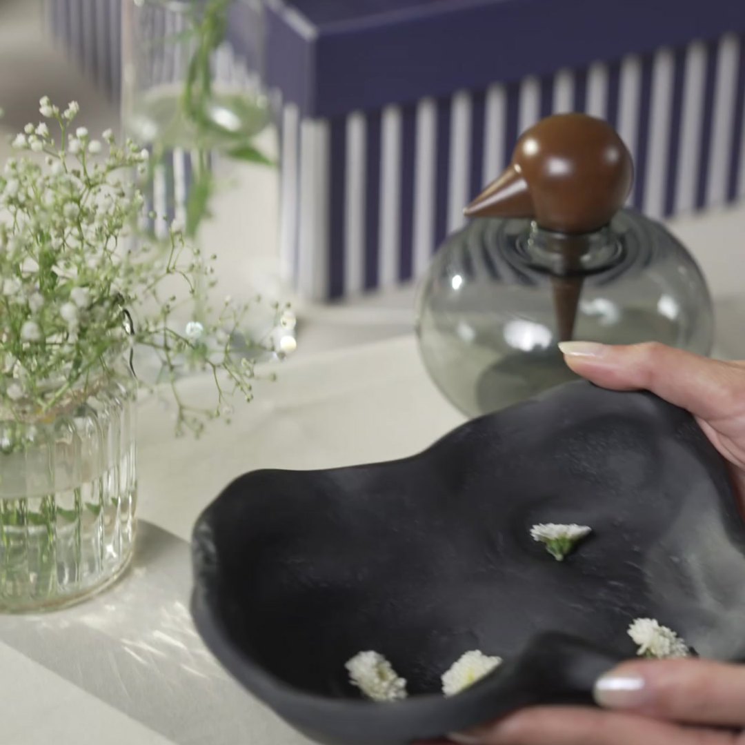 A hand holds a black organic-shaped ceramic kai bowl with Chirp Glass Jar next to a navy striped gift box in the background.