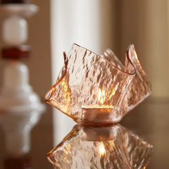 A textured peach glass candle holder with a small tea light candle inside, set on a reflective surface with a warm, softly blurred background.