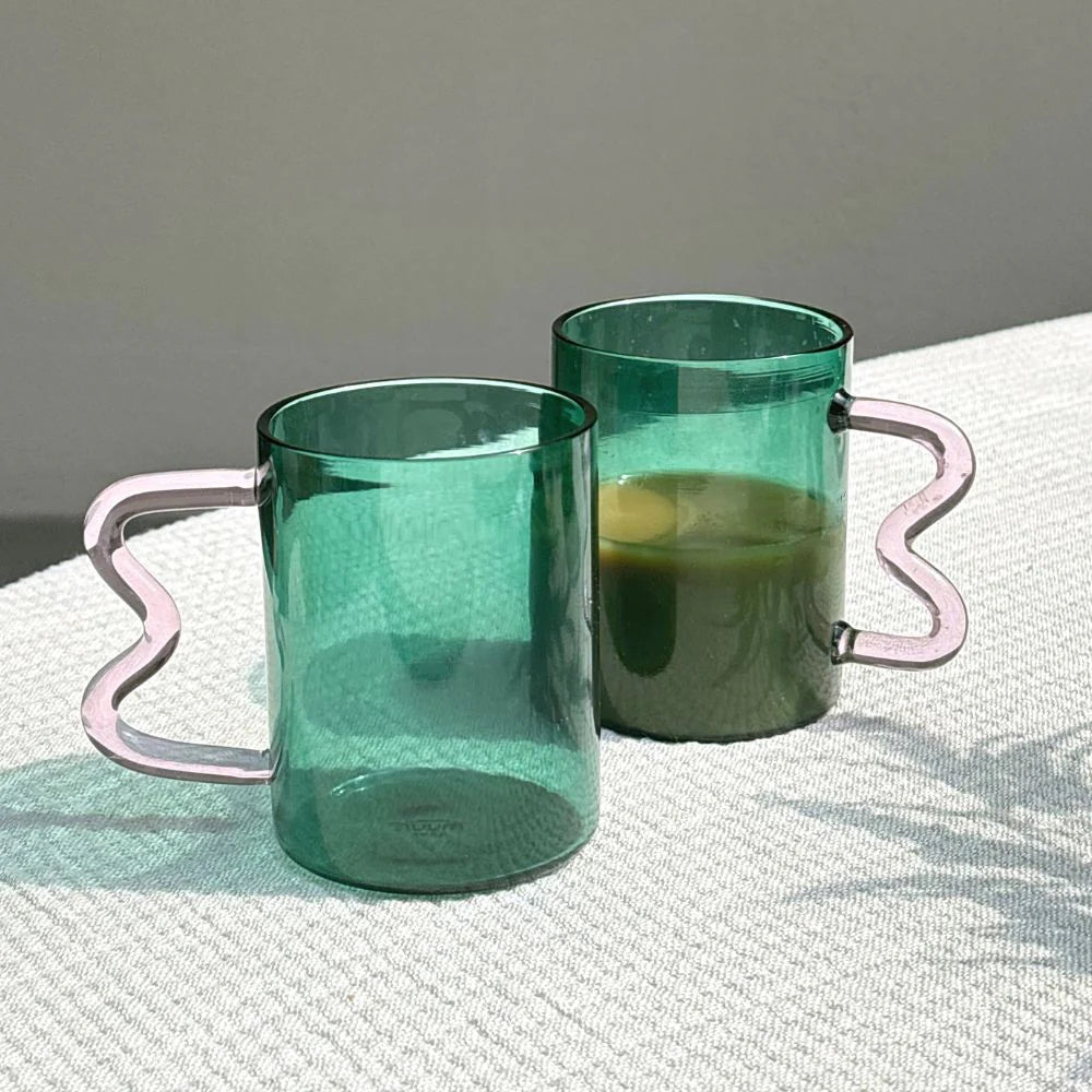 Two Swirl glass cups with teal bodies and pink handles, one filled with a dark liquid, placed on a surface with a neutral background.
