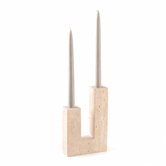 A modern U-shaped candle holder made of light beige travertine stone, holding two slim gray taper candles, set against a plain white background.