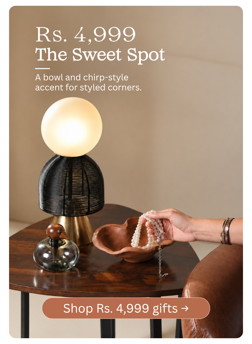 A styled interior scene showing a table lamp with a glowing spherical shade, a black ribbed base, and a gold-toned footed pedestal. Along with a Chirp Glass Jar & Kai wooden bowl