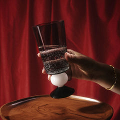 A person's hand holding a clear glass twilight tumbler, which appears to be made of borosilicate glass, set against a red background.