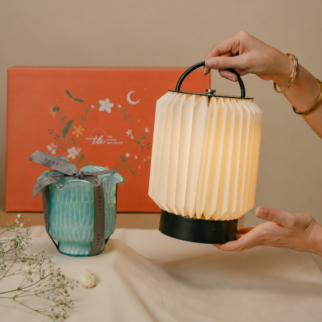 A person holds a pleated tote Portable Table Lamp with a black handle and base, illuminated from within. A teal Flower Cut Votive Candle with a gray ribbon sits on the table beside it, with a large orange floral box in the background.