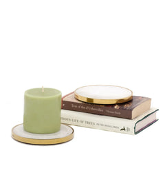 A light sage green pillar candle on a white marble Yang Candle Plate with a gold rim, styled next to a stack of three books on a white background.