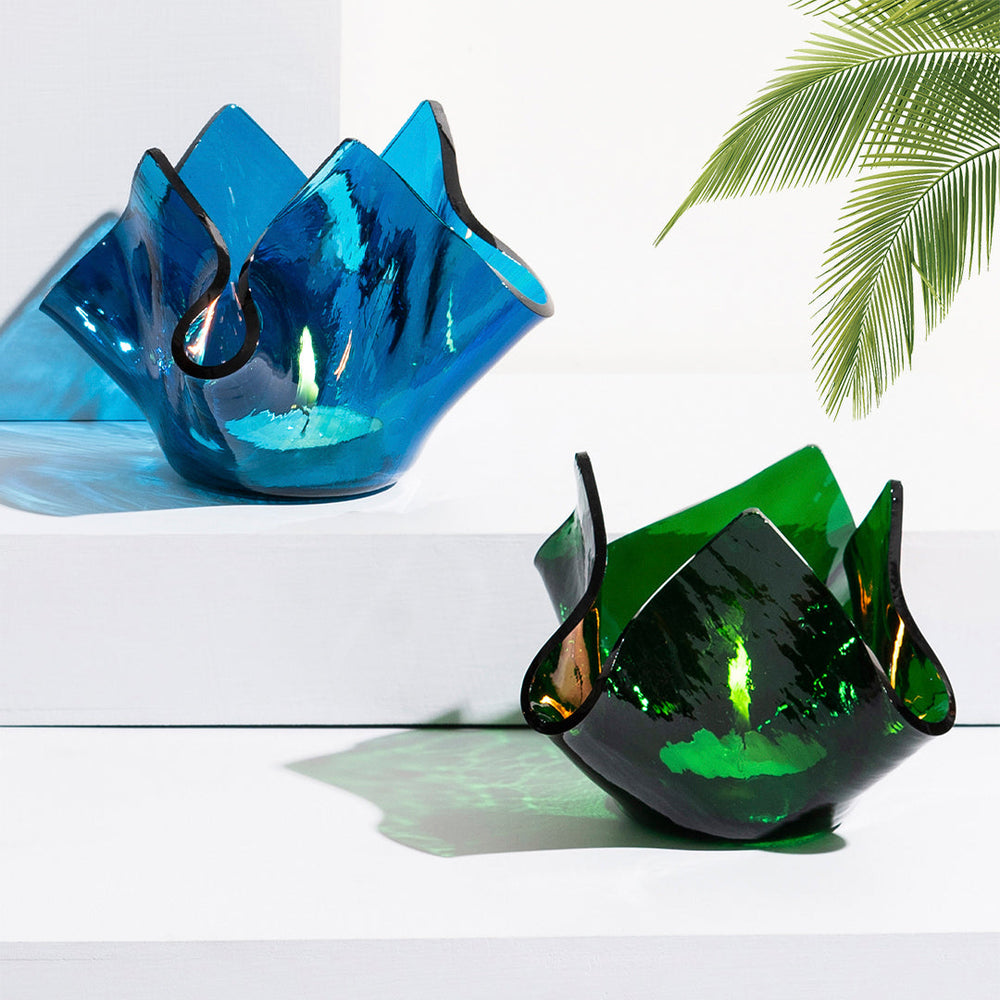 Two glass candle holders, one blue and one green, on a white surface with a palm leaf in the background.