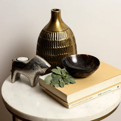 A styled interior scene showing a ribbed metallic gold Vasiliki Vase, a sculptural silver candle holder, a dark brown bowl, and two stacked beige books.
