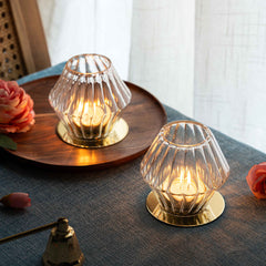 Kandeel Votive - Two glass candle holders with gold metal bases on a textured surface with flowers.