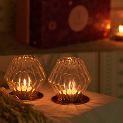 Two lit candles in decorative glass holders in diamond shaped with gold plated metal plates on a table with a blurred background