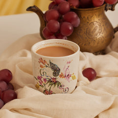 A fine bone china cup with a hand-painted design of a bird and flora, adorned with 24K gold work, placed on a linen fabric. There are grapes and a bronze teapot with gold work in the background.