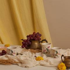Three Kulhad Botanical Affaire cups made of fine bone china with a hand-painted design of a bird and flora, placed on a table with other accessories.