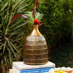 A ribbed metallic bronze ceramic vase with a gold-toned neck, holding green and burgundy foliage with a small red flower, displayed on stacked books against a blurred green garden background.