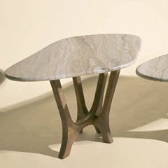 A modern accent maya table featuring an organic-shaped light gray marble top and a sculptural dark brown wooden base with three curved legs, set against a warm beige background.