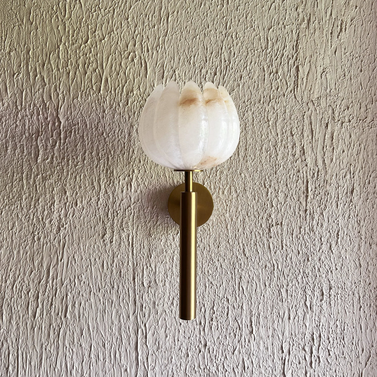 Lotus Alabaster Wall Sconce with a textured white glass flower-shaped shade and a brushed brass cylindrical stem, mounted on a textured beige plaster wall.