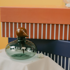 A green-tinted glass bottle with a sculptural duck-shaped wooden stopper, displayed on a white draped surface in front of colorful striped gift boxes.