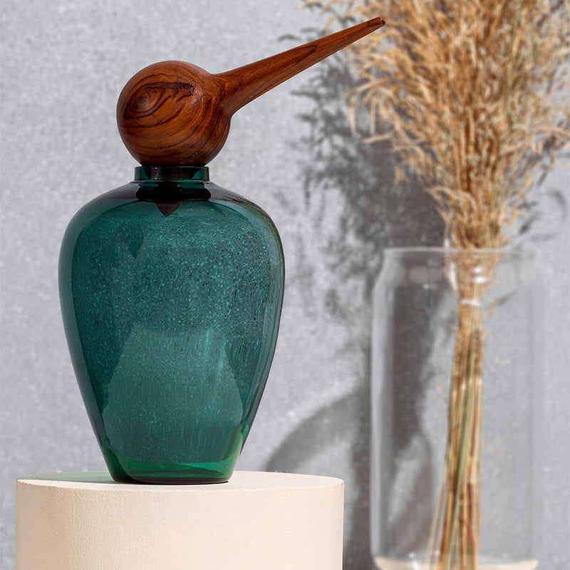 Chirp Teal glass bottle with wooden nozzle on a white pedestal against a gray background with dried plants.