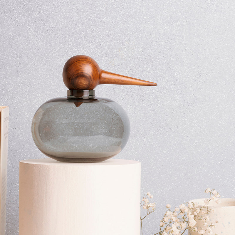 A gray hand-blown Borosilicate glass jar with a bird-shaped top on a pedestal, with a wooden beak and eye detail.