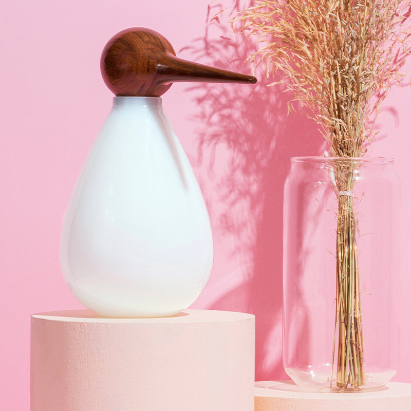 A white hand-blown glass jar shaped like a bird with a wooden beak, displayed on a pink background alongside a clear vase with dried plants.