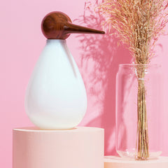 A white hand-blown glass jar shaped like a bird with a wooden beak, displayed on a pink background alongside a clear vase with dried plants.