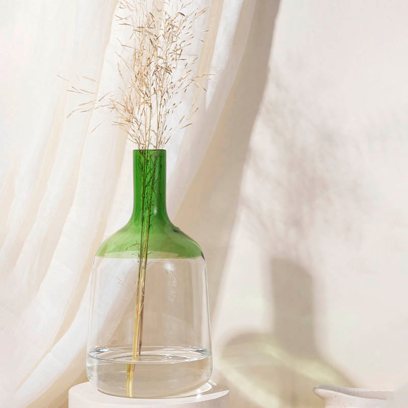 A medium-sized green glass Iris Vase with a solid pattern, containing a stalk of yellow flowers, placed on a white surface next to a white curtain.