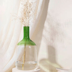 A medium-sized green glass Iris Vase with a solid pattern, containing a stalk of yellow flowers, placed on a white surface next to a white curtain.
