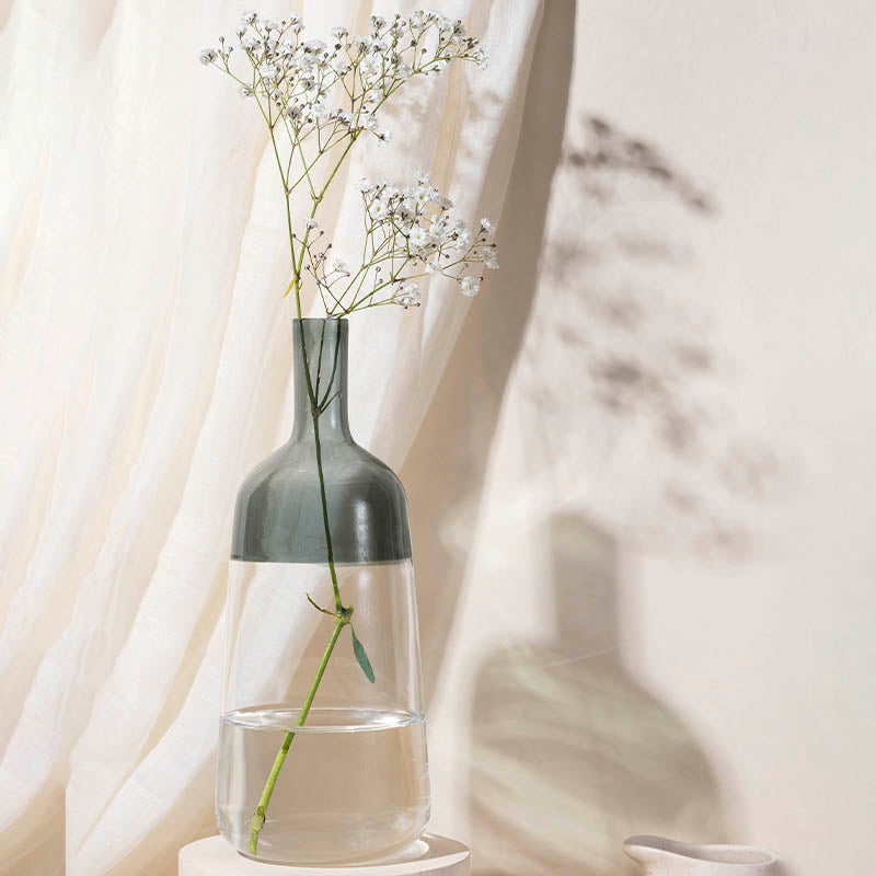 A two-tone gray glass vase with a clear base, containing flowers, placed next to a curtain on a light background.