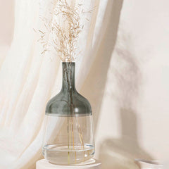 A medium-sized grey glass vase with a clear lower half, featuring a narrow neck and bell-shaped body, displayed with some dry flowers inside against a draped white background.