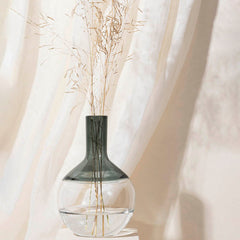 A grey glass vase with a transparent base, containing dried flowers, placed on a surface with a light curtain background.