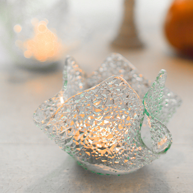 A clear glass votive holder with a crumpled texture design, illuminated by a candle inside, placed on a wooden surface.