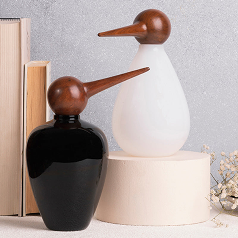 Two bird-shaped hand-blown glass jars in black and white colors, standing one inside the other on a beige surface, with a book and a vase in the background.
