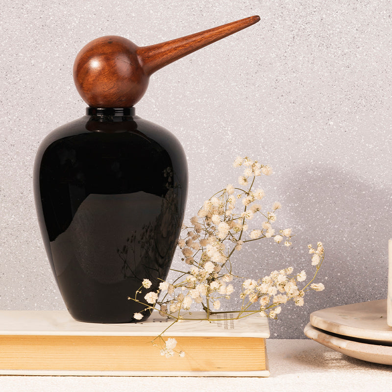A black glass jar with a bird-shaped top, placed on a table with a few white flowers beside it.