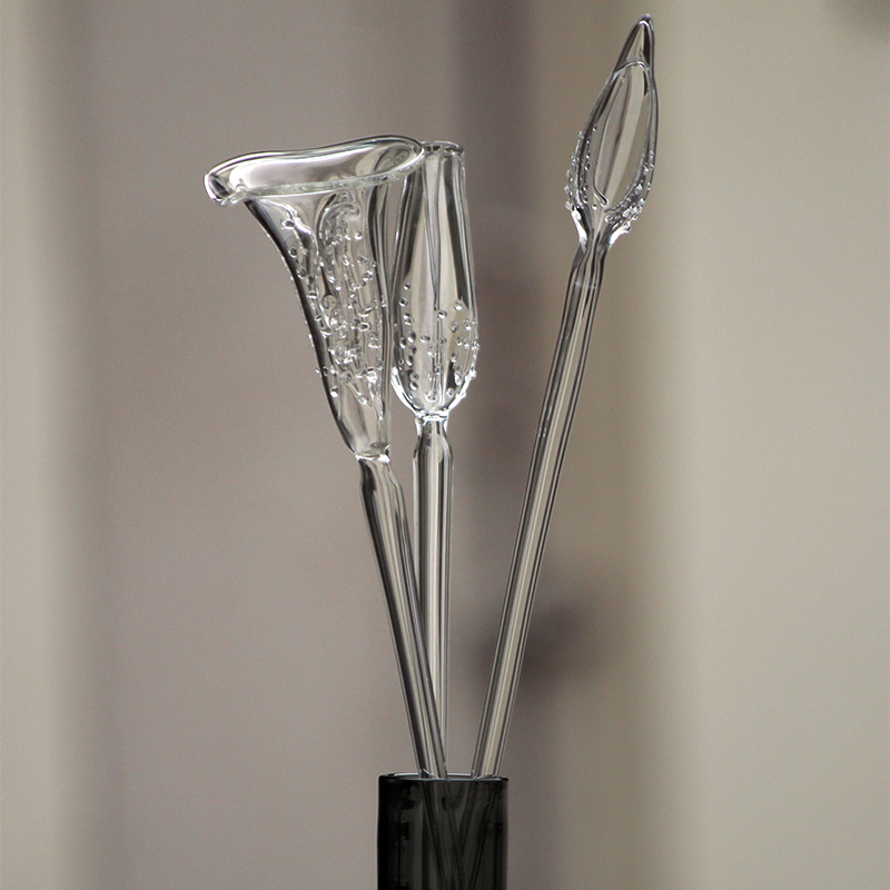 Three clear calla-lily-shaped decorative glass stems arranged in a dark vase against a softly blurred warm neutral background.