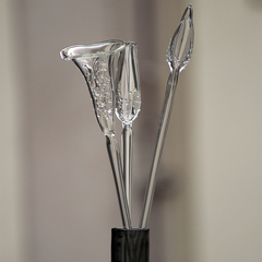 Three clear calla-lily-shaped decorative glass stems arranged in a dark vase against a softly blurred warm neutral background.