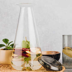 Iris Glass Carafe, made of borosilicate clear glass with grey borosilicate glass lid placed next to it on a table with glass, bowl and small plant 