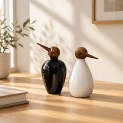 Two decorative penguin-shaped glass bottles with wooden beaks and heads, one glossy black and one matte white, displayed on a wooden table in a softly lit room.