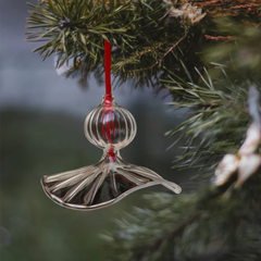 A clear, fluted handblown glass ornament shaped like a flower, hanging from a red ribbon on a Christmas tree.