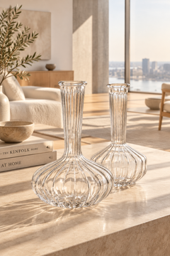 Two clear ribbed glass decanters with bulb-shaped bases and narrow flared necks, displayed on a light stone table in a sunlit modern living room.