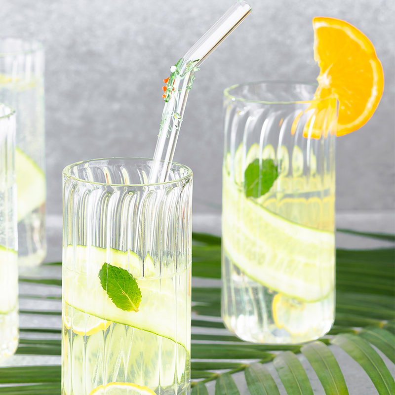 Two tall clear firdaus cocktail glasses with a ribbed/fluted design, stem-less. One glass has a slice of orange and other glass of straw. 