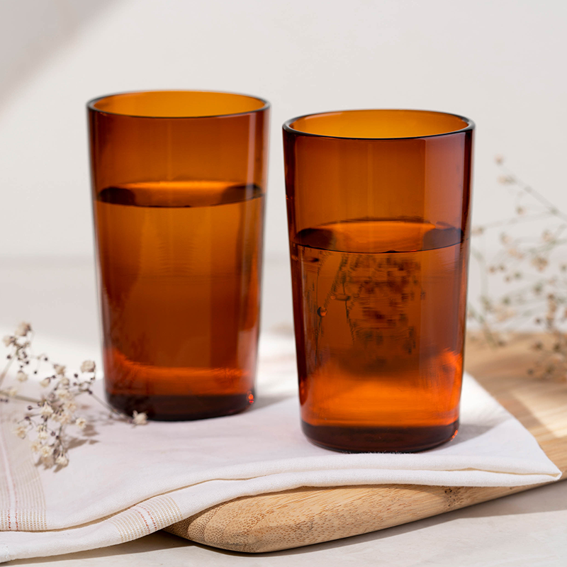 Two orange colored Juliette glass highball glasses on a white napkin, with a wooden board and some dried flowers in the background.