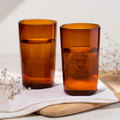 Two orange colored Juliette glass highball glasses on a white napkin, with a wooden board and some dried flowers in the background.