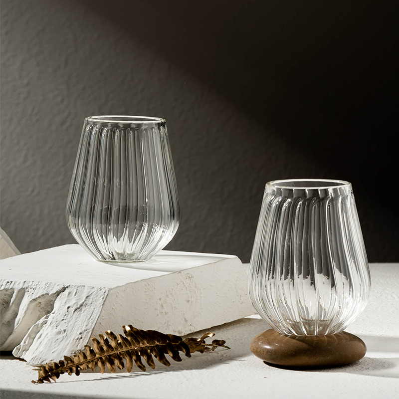 Two clear stemless glasses with a straight ribbed design, placed on a white surface, with a wooden base visible on one glass.