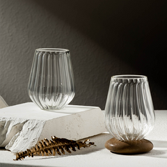 Two clear stemless glasses with a straight ribbed design, placed on a white surface, with a wooden base visible on one glass.