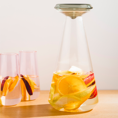 A clear glass Iris carafe with a glass lid, filled with what appears to be water and fruit slices, placed on a table with a window in the background.