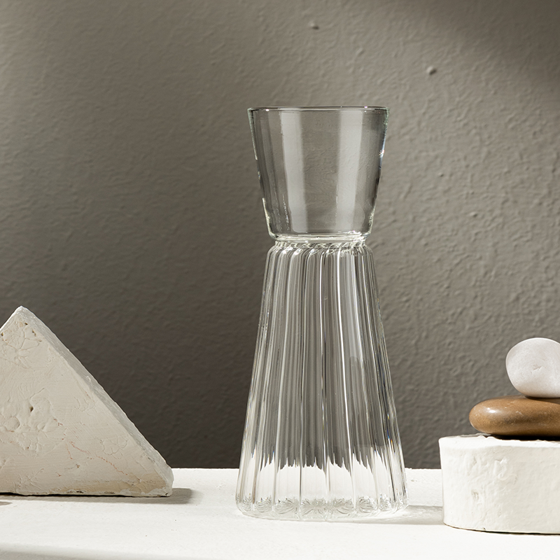 A clear Firdaus glass carafe with a fluted pattern, displayed next to a white geometric shape and a marble-like object on a light surface.