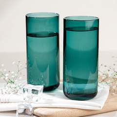 Two teal colored Juliette cocktail glasses on a wooden surface with a white napkin and ice cubes, garnished with a sprig of herbs.