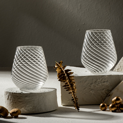 Two clear ribbed stemless glasses with a twisted design, standing on white foam blocks, with a dry branch and golden nuts nearby.